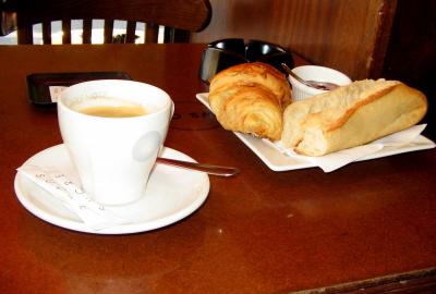 La imagen muestra una mesa de bar sobre la cual hay un café, una croissant, un pan y mermelada.