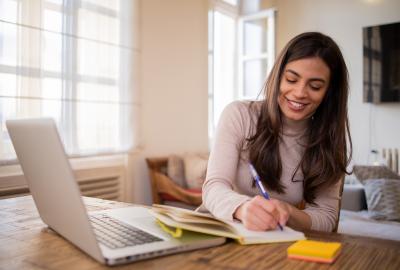 La imagen muestra a una mujer sonriendo y estudiando en su computadora, anotando en un cuaderno