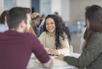 La imagen muestra a un grupo de personas charlando en un aula, una mujer con pelo ondulado sonríe