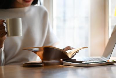 La imagen muestra una mujer frente a una computadora, también tiene una taza en la mano y con la otra sostiene un libro