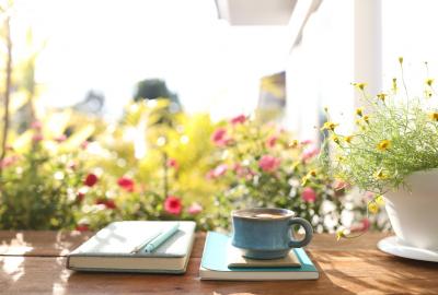 La imagen muestra un escritorio mirando al exterior, sobre el cual se apoyan un libro, un cuaderno y una taza de café