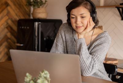 La imagen muestra a una mujer con auriculares mirando una notebook, está en su cocina