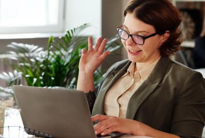 La imagen muestra a una mujer frente a una computadora saludando hacia cámara