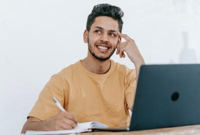 En la imagen se ve a un joven muchacho, de cabello y barba castaños, está sonriendo y pensativo frente a una computadora. También tiene una lapicera en la mano y anota algo en un cuaderno.