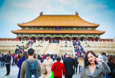 En la imagen se ve a una mujer china mirando a cámara y sonriendo, a sus espaldas se ve un templo oriental de la CIudad Prohibida