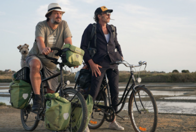 La imagen muestra un fotograma de la película À bicyclette, de Mathias Mlekuz. Dos hombres en bicicleta a orillas de un campo. 