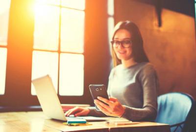 En la imagen se ve una muchacha sentada frente a una computadora, mirando un celular y sonriendo. Al lado una ventana por donde se ve el sol.
