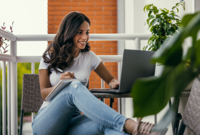 La imagen muestra a una mujer joven sentada en el balcón de su casa rodeada de plantas mirando una computadora y sonriendo